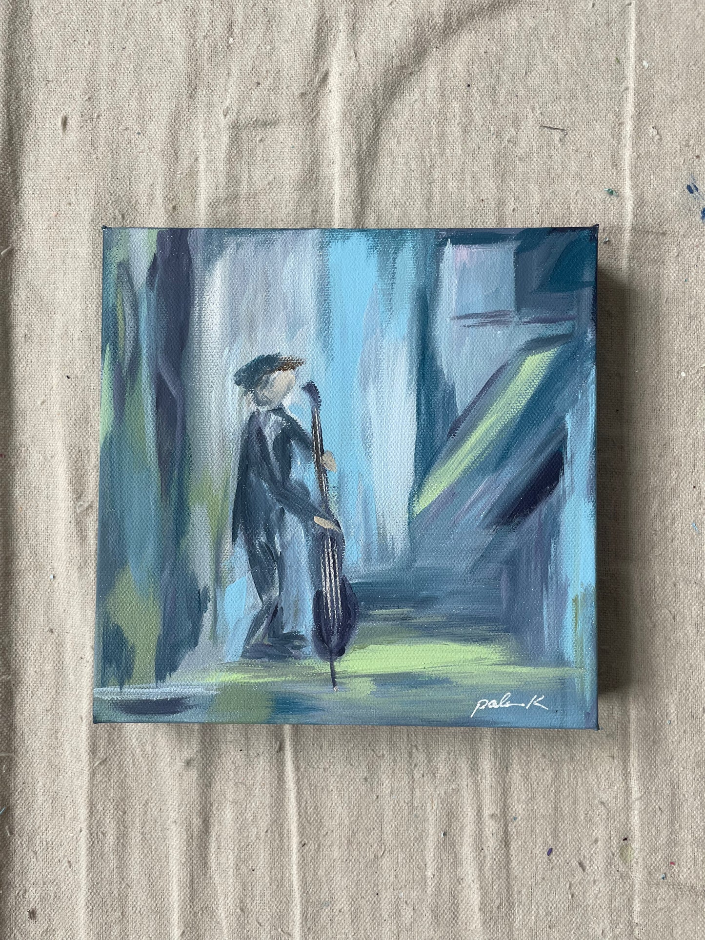 A small square canvas painting depicting an abstract figure in motion, rendered in shades of blue, gray, black, and pale yellow-green, displayed on a textured beige fabric background.